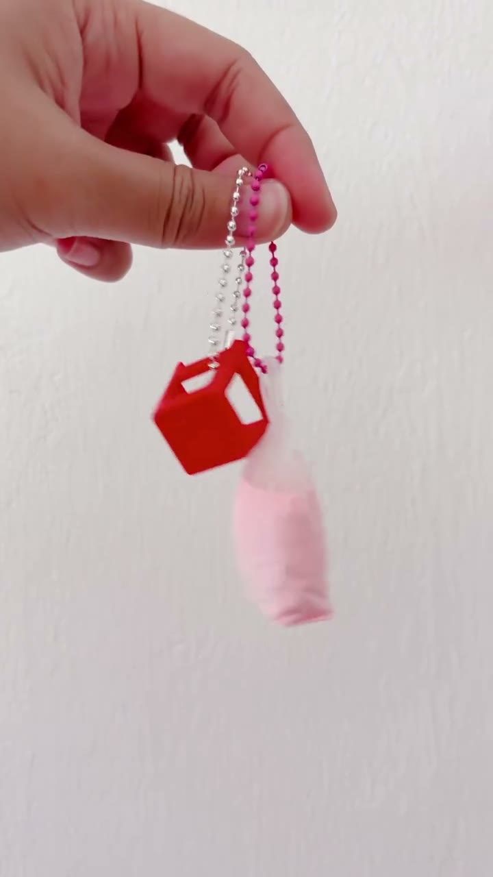 A red kenduri stool keychain and a pink tied drink bag keychain held up on ball chains